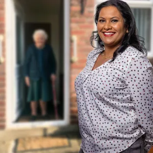 Een zorgmedewerker van ViVa! Zorggroep glimlacht vriendelijk naar de camera terwijl een cliënt, die op de achtergrond staat met een wandelstok, in de deuropening van haar woning staat.