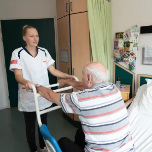 Een zorgmedewerker ondersteunt een cliënt in bed door hem te helpen staan met een hulpmiddel, in een lichte en zorgzame ruimte.