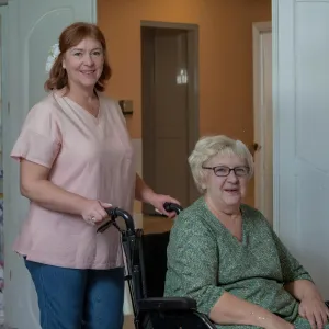 Een medewerker en een cliënt, beiden lachend, staan in een lichte gang, met de medewerker die de rolstoel van de cliënt begeleidt bij ViVa! Zorggroep