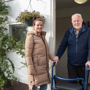 Een zorgmedewerker helpt een cliënt met een looprek voor de deur van zijn woning, bij ViVa! Zorggroep.