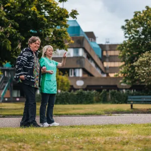 Cliënt en medewerker wandelen buiten met woonzorglocatie van ViVa! op de achtergrond