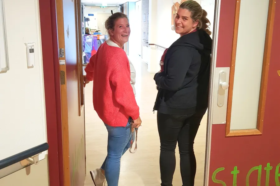 Twee zorgmedewerkers lachen naar de camera terwijl ze zich omdraaien in een gang bij ViVa! Zorggroep, klaar om hun dag te beginnen.