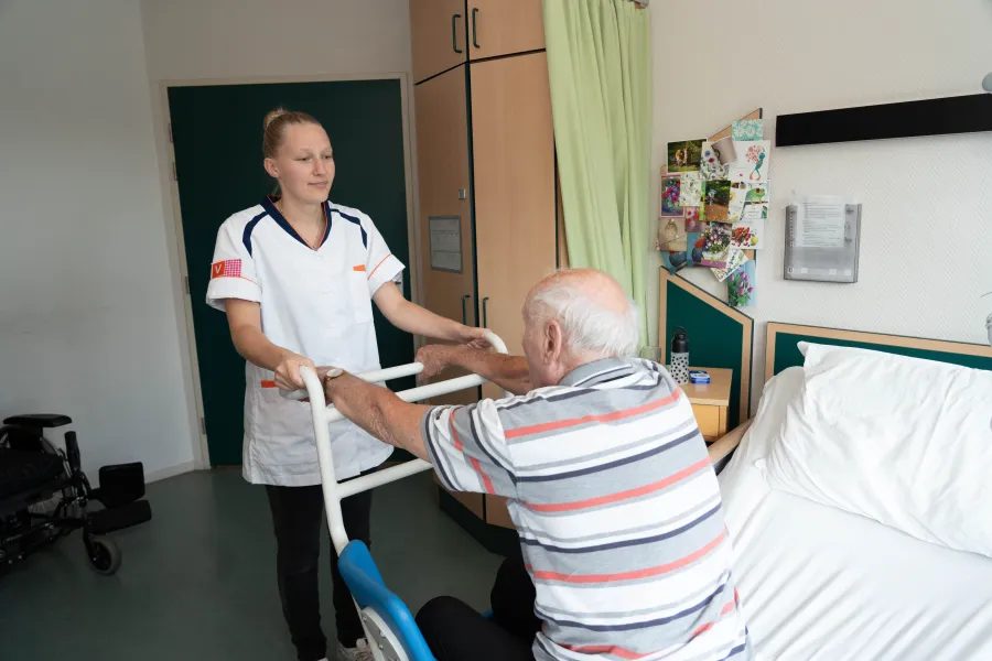 Een zorgmedewerker ondersteunt een cliënt in bed door hem te helpen staan met een hulpmiddel, in een lichte en zorgzame ruimte.
