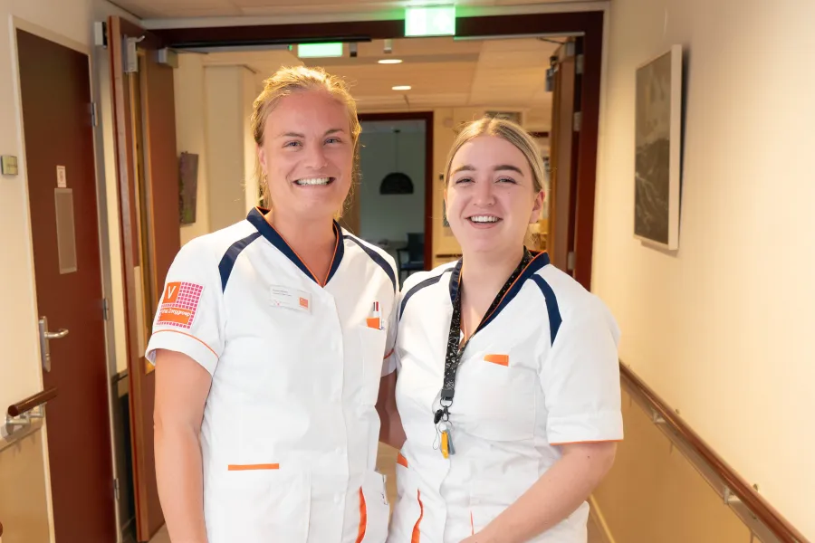 Twee medewerkers in zorgkleding staan samen in een gang bij ViVa! Zorggroep, beiden met een glimlach op hun gezicht.