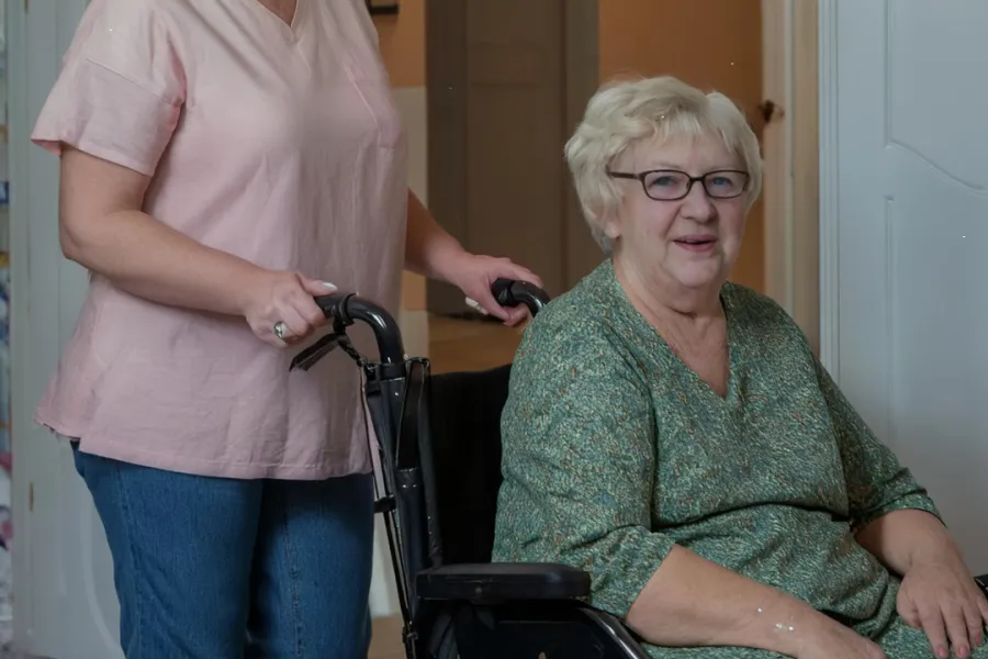 Een medewerker en een cliënt, beiden lachend, staan in een lichte gang, met de medewerker die de rolstoel van de cliënt begeleidt bij ViVa! Zorggroep