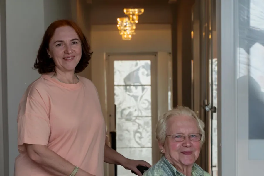 Een medewerker en een cliënt, beiden lachend, staan in een lichte gang, met de medewerker die de rolstoel van de cliënt begeleidt bij ViVa! Zorggroep