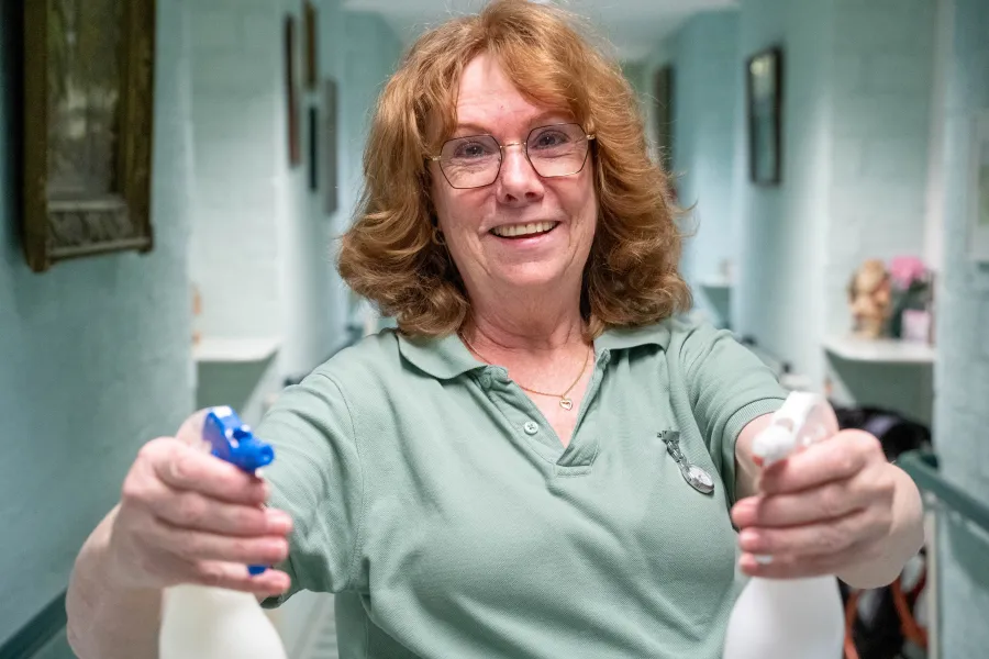 Een medewerker van ViVa! Zorggroep glimlacht terwijl ze klaarstaat met twee sprayflessen, klaar om de ruimte schoon te maken.