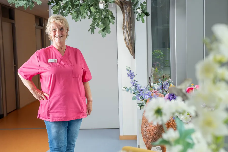 Een zorgmedewerker van ViVa! Zorggroep staat trots voor een bloeiende ruimte, gekleed in het roze uniform, met een glimlach die vertrouwen en zorg uitstraalt.