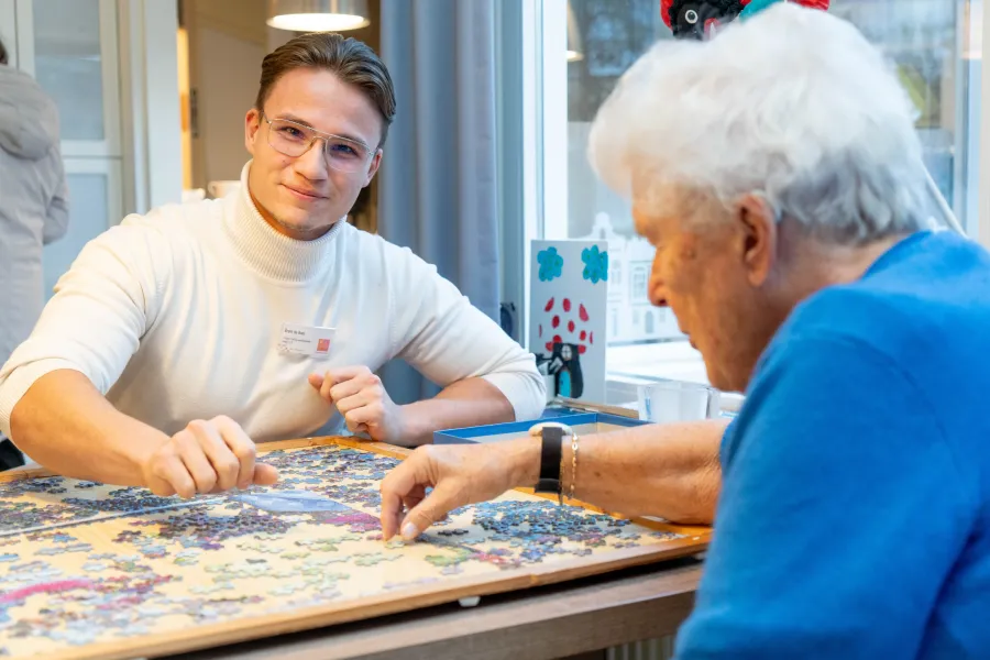 Een medewerker van ViVa! Zorggroep en een cliënt genieten samen van een moment van ontspanning met een puzzel in een sfeervolle woonkamer.
