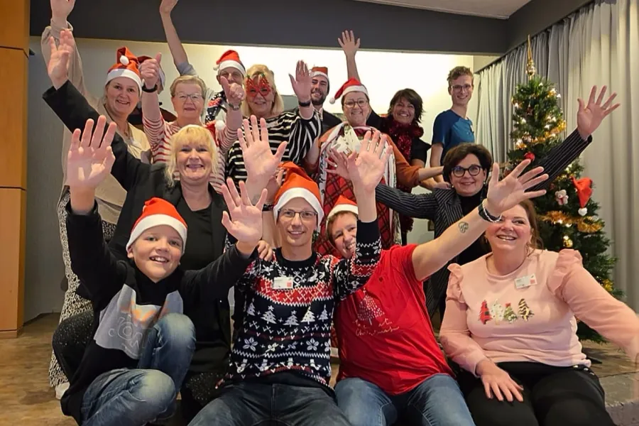 Een vrolijke groep ViVa!-collega’s viert samen de feestdagen. Iedereen draagt een kerstmuts, lacht en zwaait naar de camera, terwijl op de achtergrond een versierde kerstboom staat. De sfeer is warm, gezellig en vol teamspirit bij ViVa! Zorggroep.